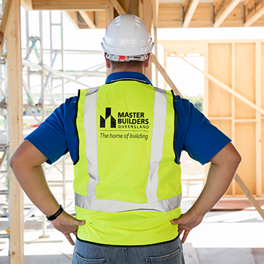 A construction worker faces away from the camera. He is wearing a white hard hat and a yellow high vis vest that has the Master Builders logo on it